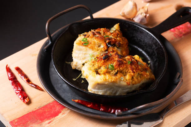 food concept spot focus homemade butter, garlic, bread crumbs and cheese crunchy baked cod fish in skillet iron cast pan on black background with copy space - pollock trawler imagens e fotografias de stock