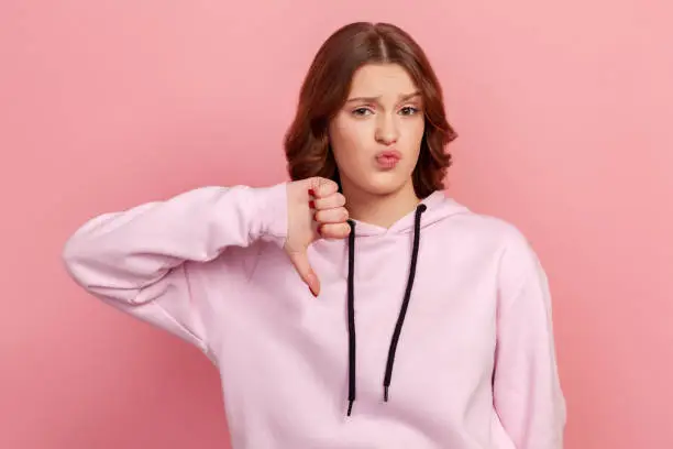 Portrait of naughty dissatisfied brunette teen in hoodie frowning angrily and showing thumbs down gesture, expressing disapproval, dislike. Indoor studio shot isolated on pink background