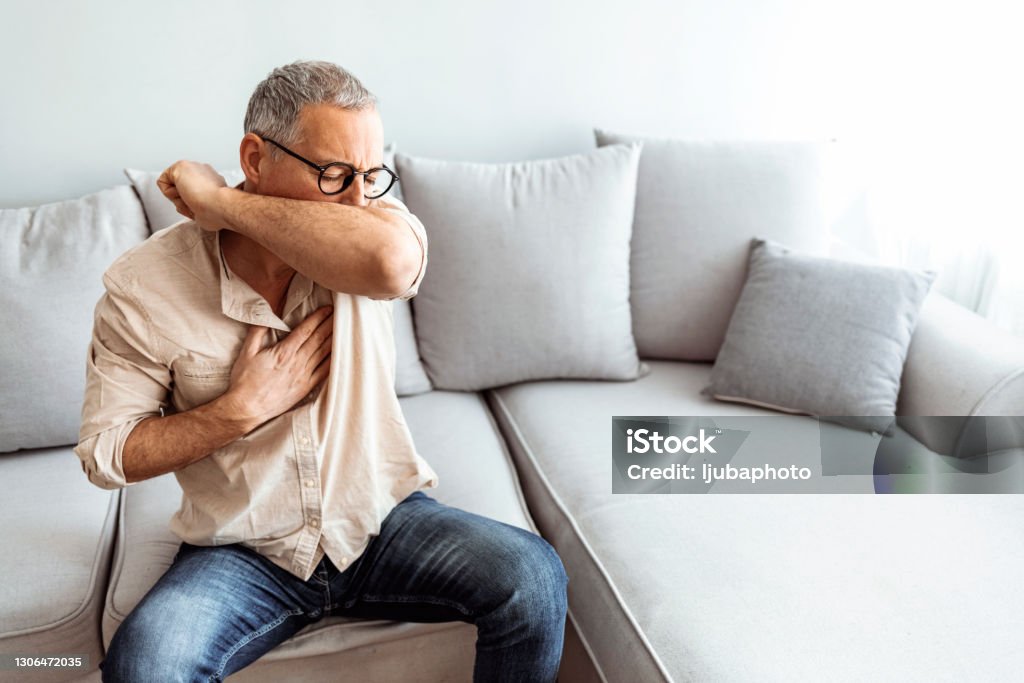Being sick on a beautiful day is the worst Caucasian male coughs in his elbow while sitting on sofa at home. Correct sneezing. Concept of stop spread of the virus. Coughing Stock Photo