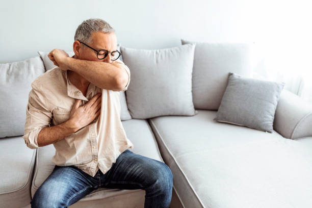 Being sick on a beautiful day is the worst Caucasian male coughs in his elbow while sitting on sofa at home. Correct sneezing. Concept of stop spread of the virus. respiratory disease stock pictures, royalty-free photos & images