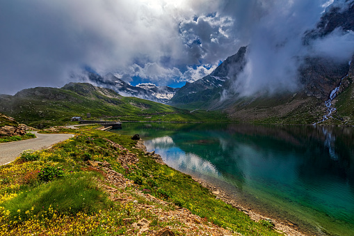 View of road running along green shores and small alpine lake under low clouds as mountains on background in Piedmont, Northern Italy.