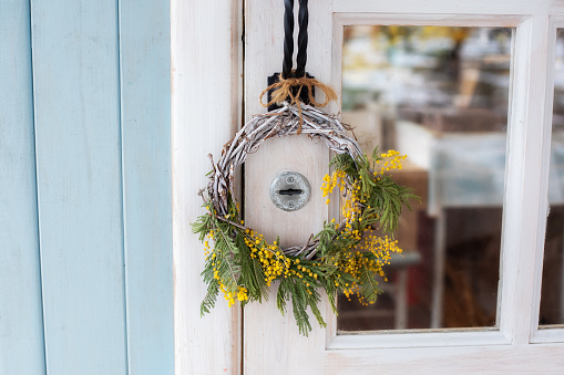 Front Door with wreath of Mimosa yellow flowers. Easter wreath. Spring decoration on wooden door of house. Home entrance with decorative Spring wreath on door. Rustic interior element of spring porch