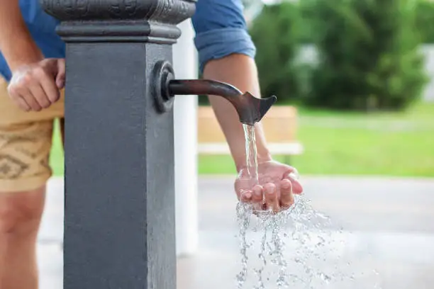 Man washing his hand in faucet water. Сity water tap with drinkable water in park. Drinking column. Ancient black column for distribution of drinking water installed on street. Watertap on hydrant