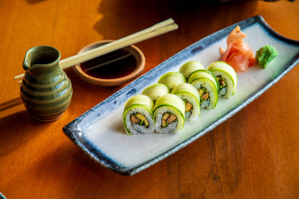 istock Cucumber coated sushi rolls served with ginger and wasabi on rectangular plate, soy sauce with chopsticks on the table 1306478550