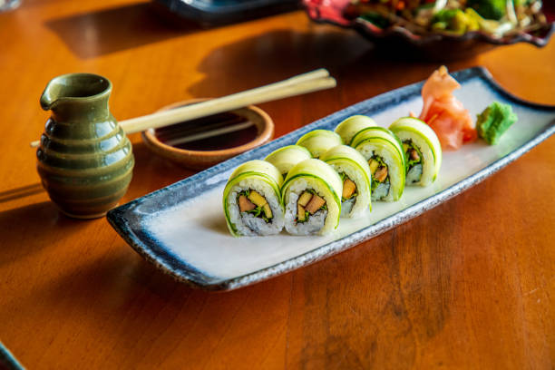 istock Cucumber coated sushi rolls served with ginger and wasabi on rectangular plate, soy sauce with chopsticks on the table 1306478562