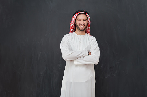 Young muslim man with crossed arms smiling. Happy Arab in traditional clothes standing in front of black chalkboard representing Ramadan Kareem concept