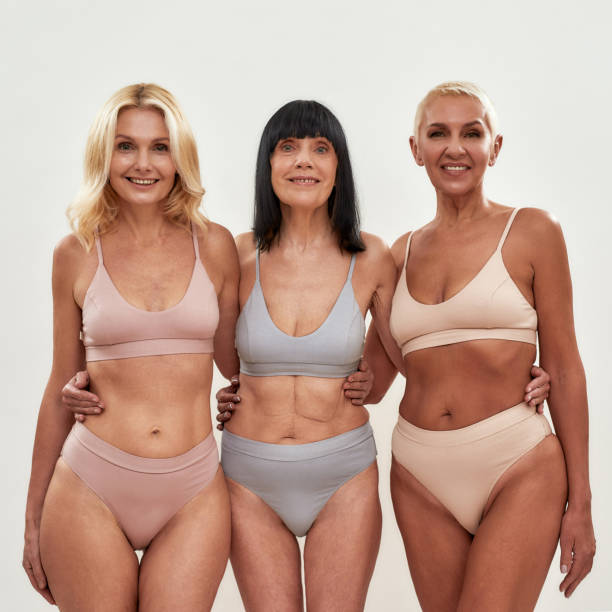 три привлекательные женщины среднего возраста в нижнем белье обнимаются, позируя вместе на светлом фоне - beauty beautiful bikini caucasian стоковые фото и изображения