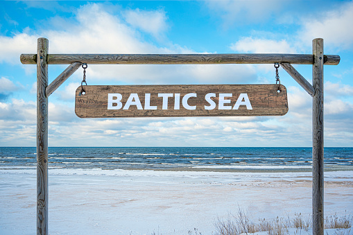 Wooden sign of Baltic sea against blue sky, sea and beach with white sand.
