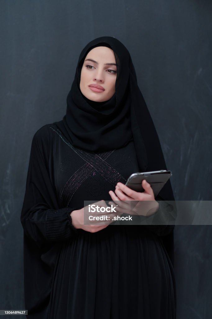 Young Arab businesswoman in traditional clothes or abaya with tablet computer Young Arab businesswoman in traditional clothes or abaya with tablet computer in front of black chalkboard representing modern islam fashion technology Abaya - Clothing Stock Photo