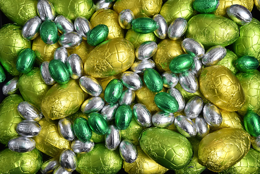 Large & small yellow, gold, green, lime green and silver spring colours of foil wrapped chocolate easter eggs, against a black background.