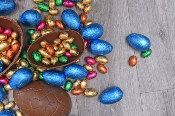 istock Pile or group of multi colored and different sizes of colourful foil wrapped chocolate easter eggs in pink, blue, gold and green. Large halves of a brown milk chocolate egg have mini eggs inside, on a grey wooden background. 1306491137