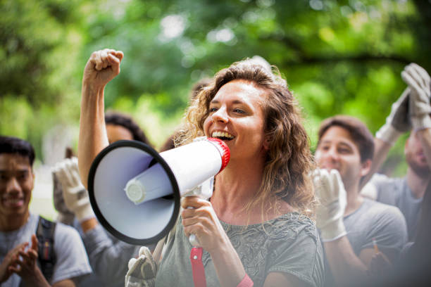 Young woman activist holding megaphone and giving a speech with her fist up during garbage cleanup event Cheerful young female volunteer talking on megaphone in park during garbage cleanup program activist stock pictures, royalty-free photos & images