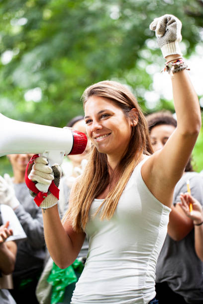 istock Young female volunteer with arm raised while talking on megaphone standing next toher friends in park 1306495456