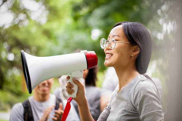Portrait of young female activist talking on megaphone during daytime Side view of smiling young female volunteer talking on megaphone in park during garbage cleanup event activist speech stock pictures, royalty-free photos & images