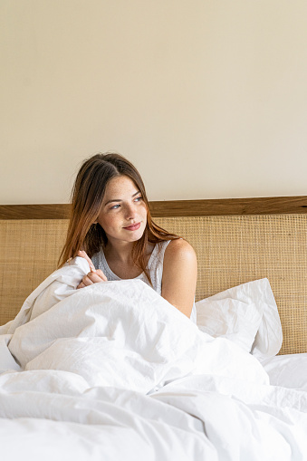 Thoughtful young woman with blanket sitting on bed at home