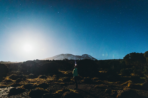 Young woman traveler walking at the beautiful terrain with view of the peak of Kilimanjaro during the night full of million of stars
