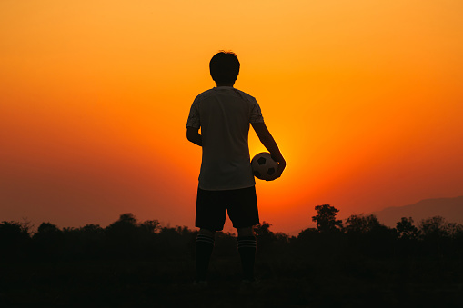 Silhouette action sport outdoors of a young man having fun playing soccer football for exercise in community rural area under the twilight sunset.