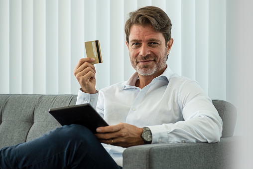 Portrait of smiling mature businessman holding credit card and digital tablet while sitting on sofa in office