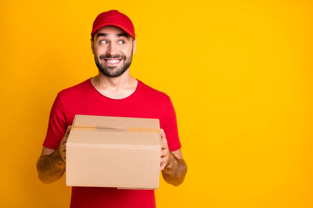 istock Photo of cute funny guy courier dressed red clothes delivering brown box looking empty space isolated yellow color background 1306516574