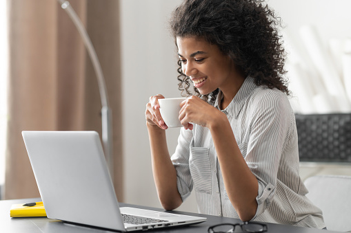 Smiling young cheerful African American woman with Afro hairstyle enjoying morning coffee while watching video news on the laptop, talking with her virtual friend, sitting at the desk in home office