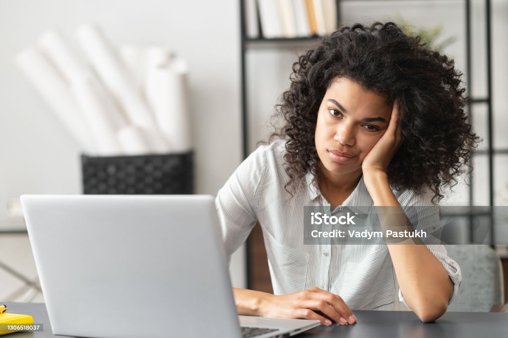 Annoyed African American woman sitting at the desk Young African American woman with afro hairstyle looks annoyed and stressed, sitting at the desk, using a laptop, thinking and looking at the camera, feeling tired and bored with depression problems Boredom Stock Photo