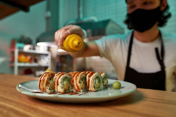 istock Professional sushi chef wearing protective mask and gloves decorating sushi rolls with falling sesame at commercial kitchen 1306518652