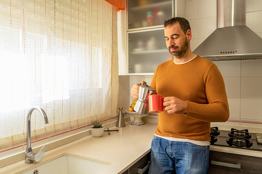 Bearded man in an orange sweater and blue jeans pouring himself a cup of coffee in the kitchen at home. Tranquility concept