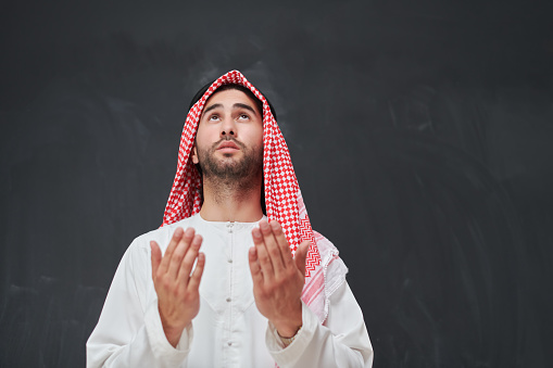 Arab man in traditional clothes praying to God or making dua. Muslim boy keeps hands in praying position in front of black chalkboard representing Ramadan concept