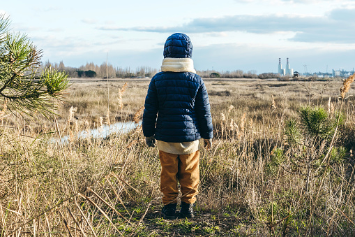 Back view of a child wearing winter clothes standing in front of wild landscape and factories on background - Activism and environmental protection concept - Childhood, future for the new generations
