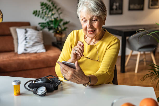 Senior woman holding a pill bottle and looking at smart phone Senior woman at home preparing to drink medical pill. She is holding a pill bottle and looking at smart phone old woman smiling tablet stock pictures, royalty-free photos & images