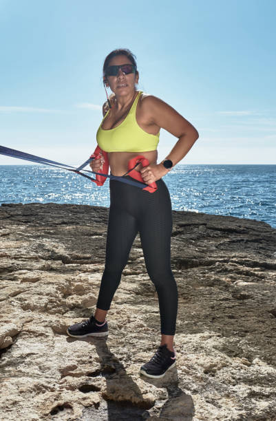 mulher latina, de meia-idade, usando roupas esportivas, treinando, fazendo exercícios físicos, prancha, abdominais, passo do alpinista, queima de calorias, manutenção de forma, ao ar livre à beira-mar, usando fones de ouvido, relógio inteligente - pilates women sit ups yoga - fotografias e filmes do acervo