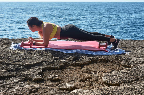 mulher latina, de meia-idade, usando roupas esportivas, treinando, fazendo exercícios físicos, prancha, abdominais, passo do alpinista, queima de calorias, manutenção de forma, ao ar livre à beira-mar, usando fones de ouvido, relógio inteligente - pilates women sit ups yoga - fotografias e filmes do acervo