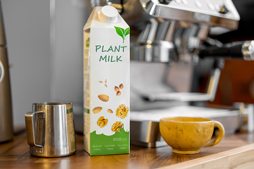 Packaging of vegetable milk near pincher, a cup of coffee and coffee machine on wooden table top on the kitchen. Organic nut dairy, alternative drink.