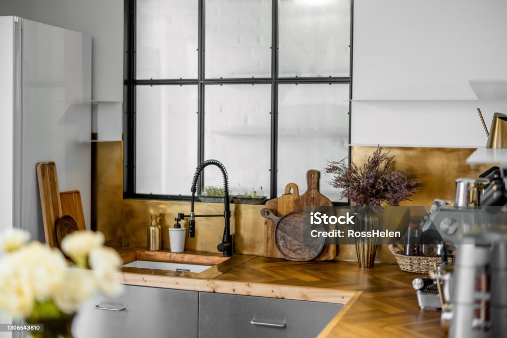 Stylish kitchen interior with wooden table top Stylish kitchen interior with wooden table top and stainless steel facades. Foreshortening of the sink area with a window with wooden boards, flowers and decor. Beauty Stock Photo