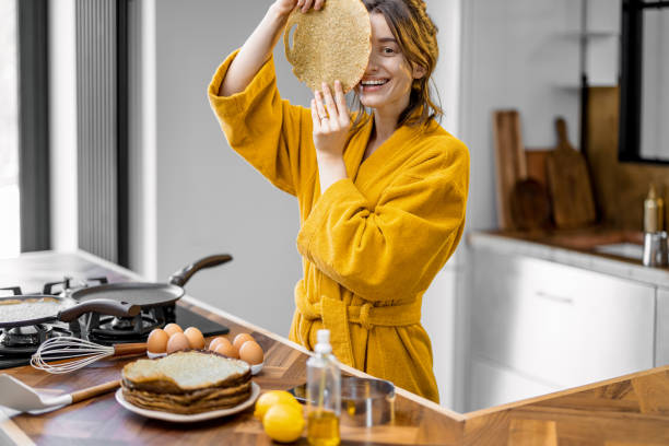 Portrait of a young joyful woman covering her face with fried pancake while cooking breakfast on the kitchen at home