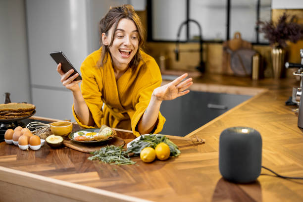 Woman speaking to a smart speaker during a breakfast at home Happy woman in bathrobe controlling home devices with a voice commands, speaking to a smart column during breakfast time on the kitchen at home. Smart home concept virtual assistant stock pictures, royalty-free photos & images