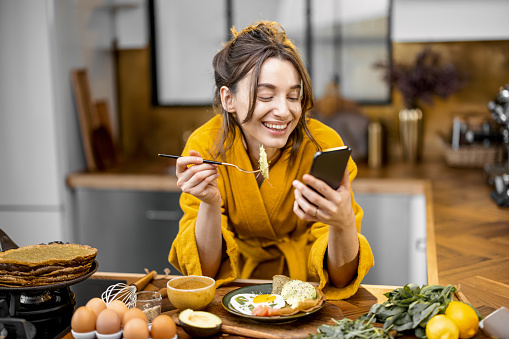 Young happy woman dressed in yellow bathrobe enjoys healthy breakfast and reading on phone at home. Morning affairs and routine