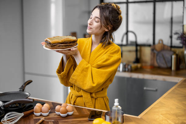 Portrait of a young cheerful housewife dressed in bathrobe with a stack of fried pancakes, preparing breakfast on the kitchen at home. Cooking healthy food and happy house chores concept