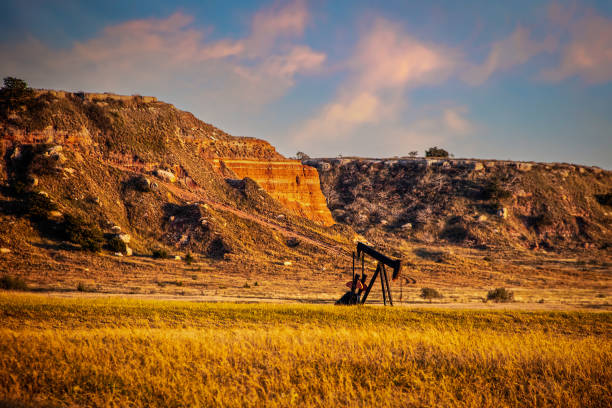 gato bomba de pozo de petróleo en tierra roja oeste de oklahoma en la hora dorada con cielo de puesta de sol temprano y acantilados en el fondo. - oil rig oklahoma oil pump oil industry fotografías e imágenes de stock