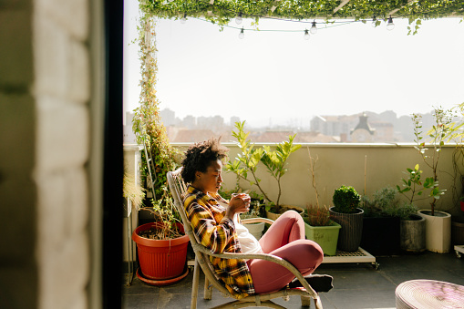 Photo of a young woman drinking her morning coffee, and enjoying the tranquility of her lovely rooftop urban garden, on a bright spring morning.