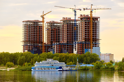 Construction site with new buildings and cranes at evening blue yellow cloudy sky background. Coast of river with  reflections water in front