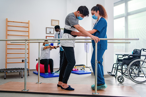 Young man using parallel bars to walk and physiotherapist helping  him  while another patient and professional are walking at the background