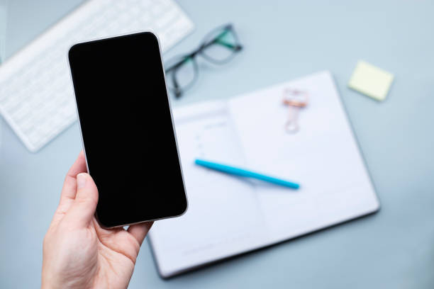 istock Smartphone in hand mock up over desktop with open planner and glasses. 1306577093