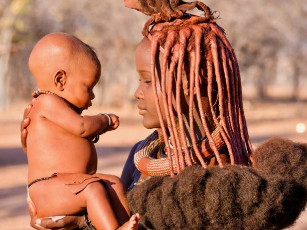 An African mother and child in a Himba village in Namibia. Rural Namibia - August 21, 2016. A young Himba woman holds her baby, while wearing the traditional tribal hairstyle and reddish skin paste made from ochre. african tribe stock pictures, royalty-free photos & images