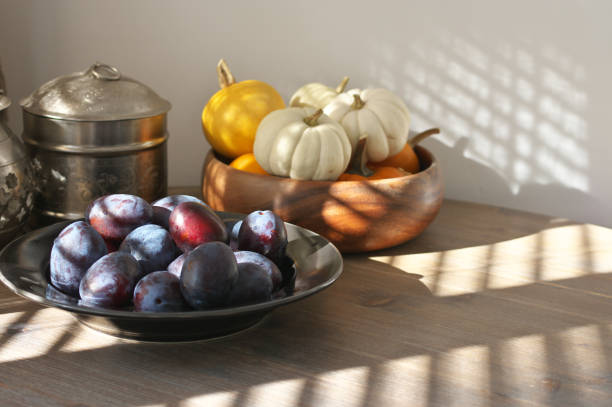 Fresh blue plums in black plate and mini pumpkins in sunlight with shadows of shutters on wood table.