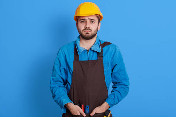 istock Helpless bearded engineer wearing yellow protective helmet keeping hands in pockets of apron, looks sad and upset, does not know what to do, posing isolated over blue background. 1306626331