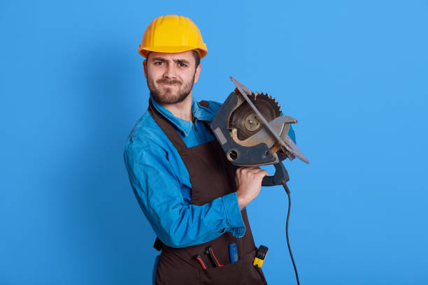 istock Strong worker with circular saw in hands looking directly at camera, frowning face, wearing protective helmet and brow apron, posing isolated over blue background. 1306626353