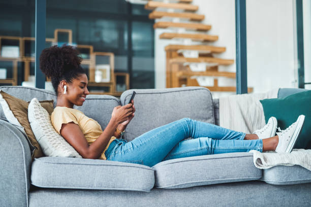 Me time is streaming time Shot of a young woman using a smartphone and wireless earbuds on the sofa at home in ear headphones stock pictures, royalty-free photos & images