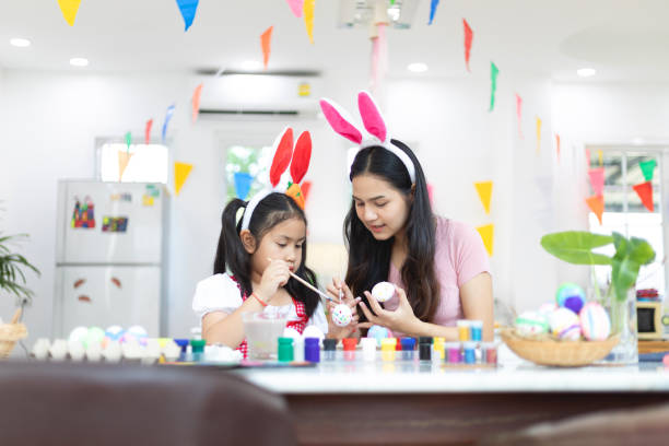 Attractive young woman with little cute girl are preparing for Easter celebration. Mom and daughter wearing bunny ears are spending time together before Easter while painting eggs. stock photo