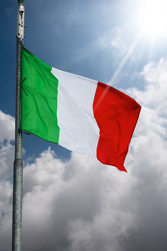 Closeup of a national Italian flag with flagpole, blowing in the wind on a blue sky with clouds and Sunbeams.